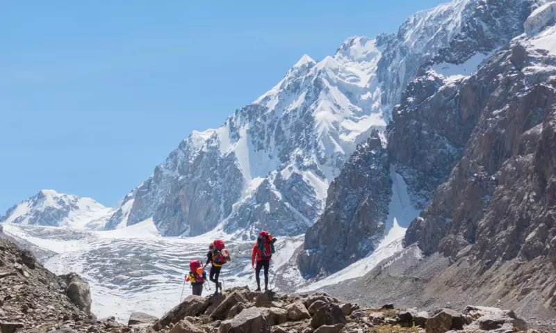 Drie wandelaars met rugzakken trekken over rotsachtig terrein richting besneeuwde bergen onder een strakblauwe hemel.